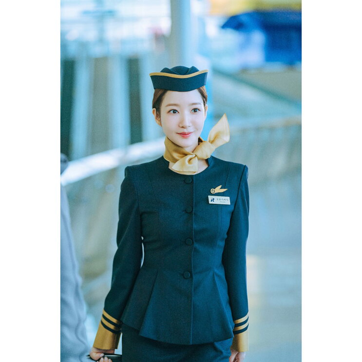 空港の屋内に、紺色の制服を着たキャビンアテンダントがいる。まとめ髪に金色の装飾が施された帽子をかぶり、金色のスカーフを巻き、胸元に羽型の社章と名札を付けている。
