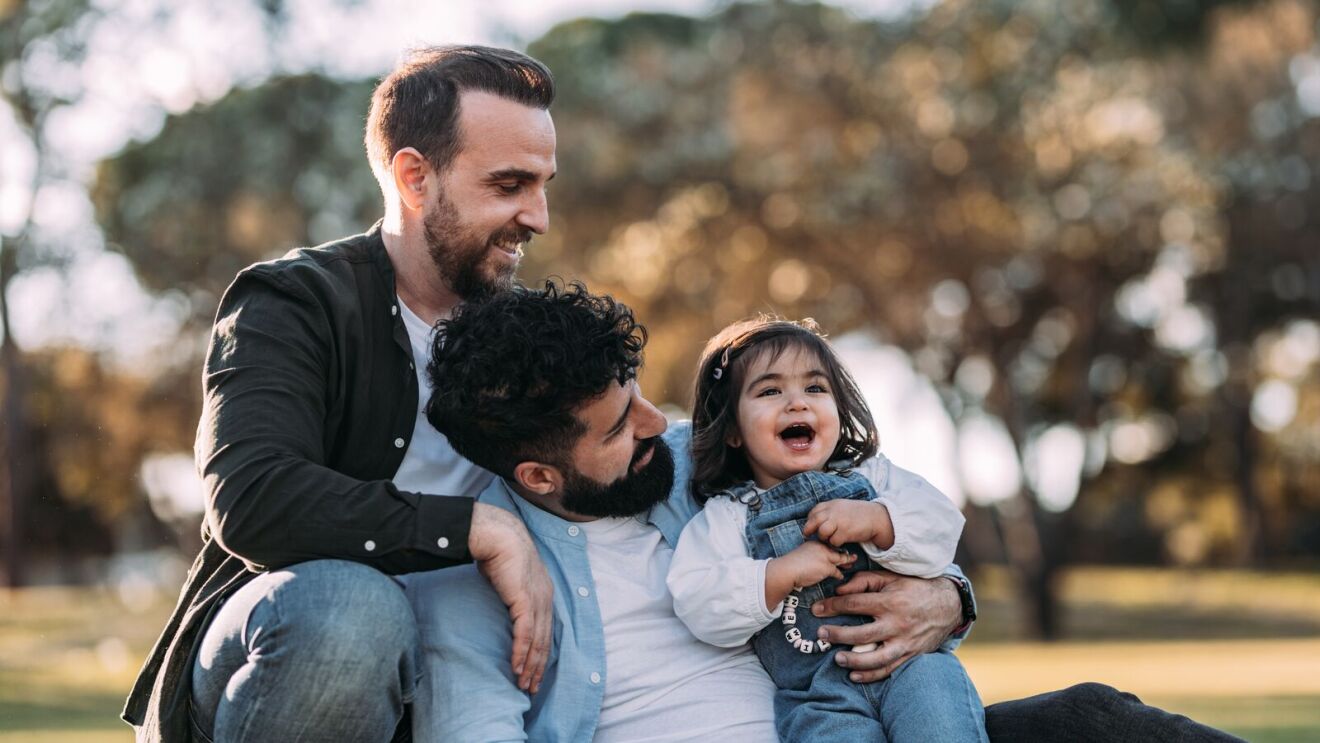 Two men and a laughing toddler embracing outdoors in a park
