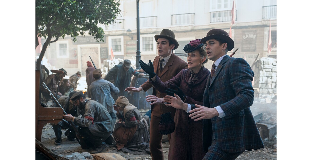 市街戦の最中と見える街中で、身なりのいい3人の人物が何かを見て両腕をあげ、驚いたような表情。3人の背後には長い銃を持ってかがんだ人々や、松葉杖をつく人物。