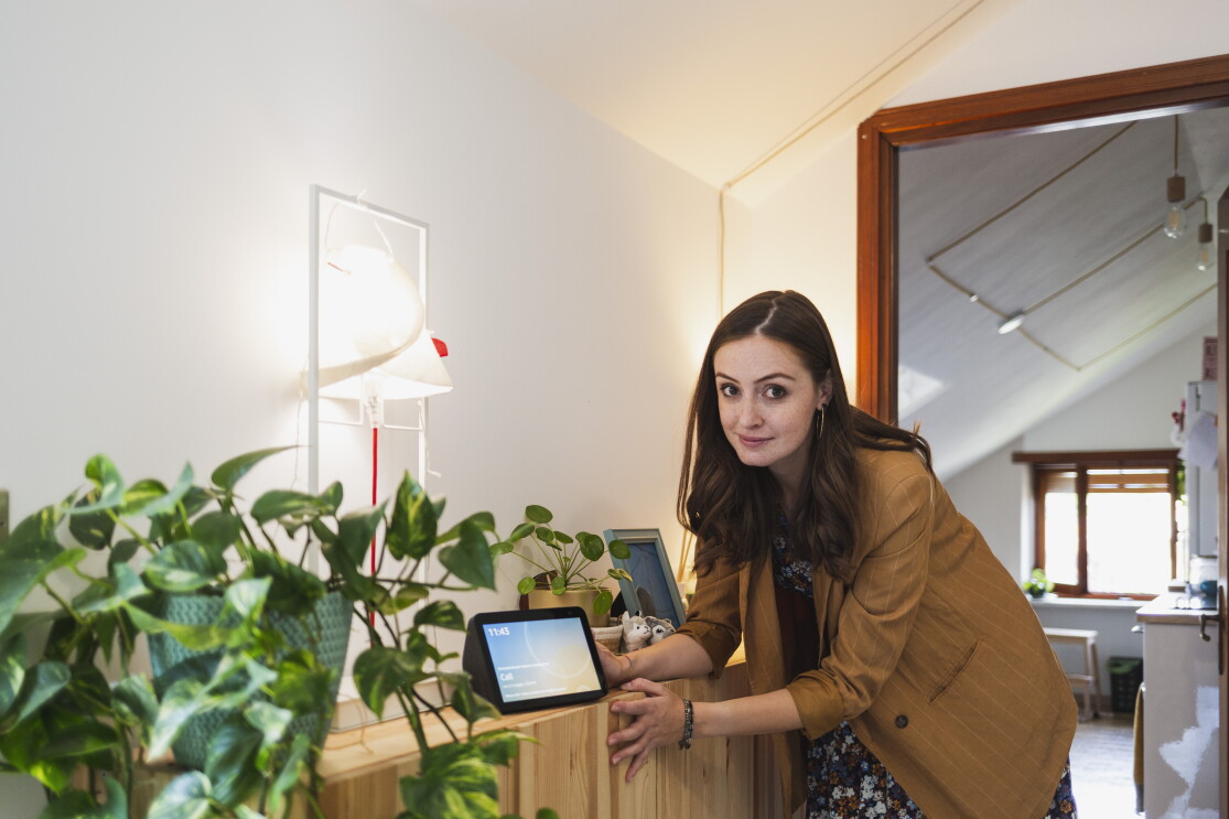 Elisa Reale, al centro della foto, è chinata verso un mobile. Guarda in camera, vicino alla mano destra ha un dispositivo Echo Show con lo schermo acceso. In primo piano, posata sullo stesso mobile, si vede una pianta in un vaso verde. Elisa indossa una giacca beige a righe bianche, ha i capelli lunghi e mossi. Sotto la giacca indossa una gonna a fiori colorata. 