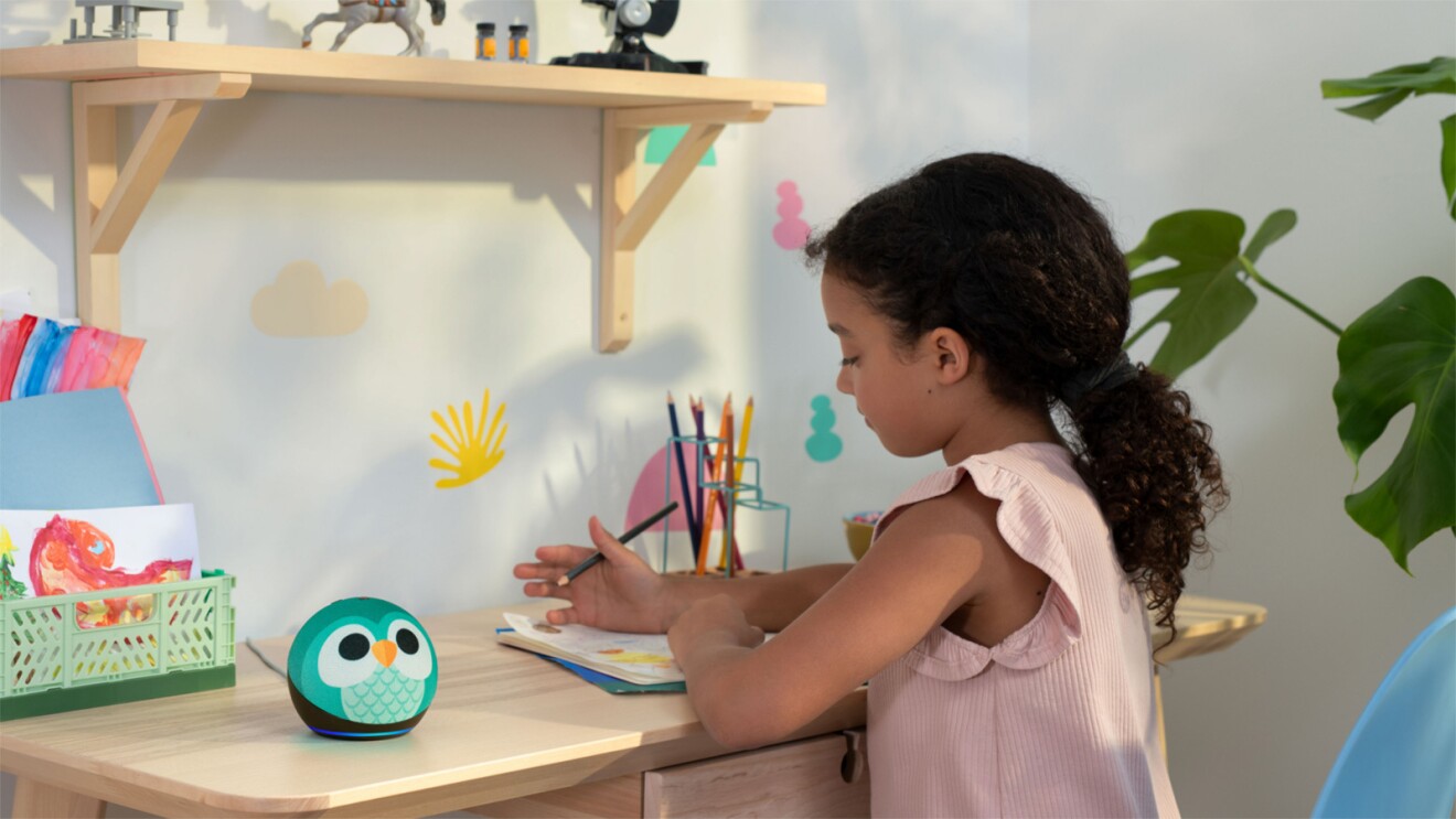 A young girl sits at a desk writing on a piece of paper. Beside her is an Echo Dot device with an owl design on it.