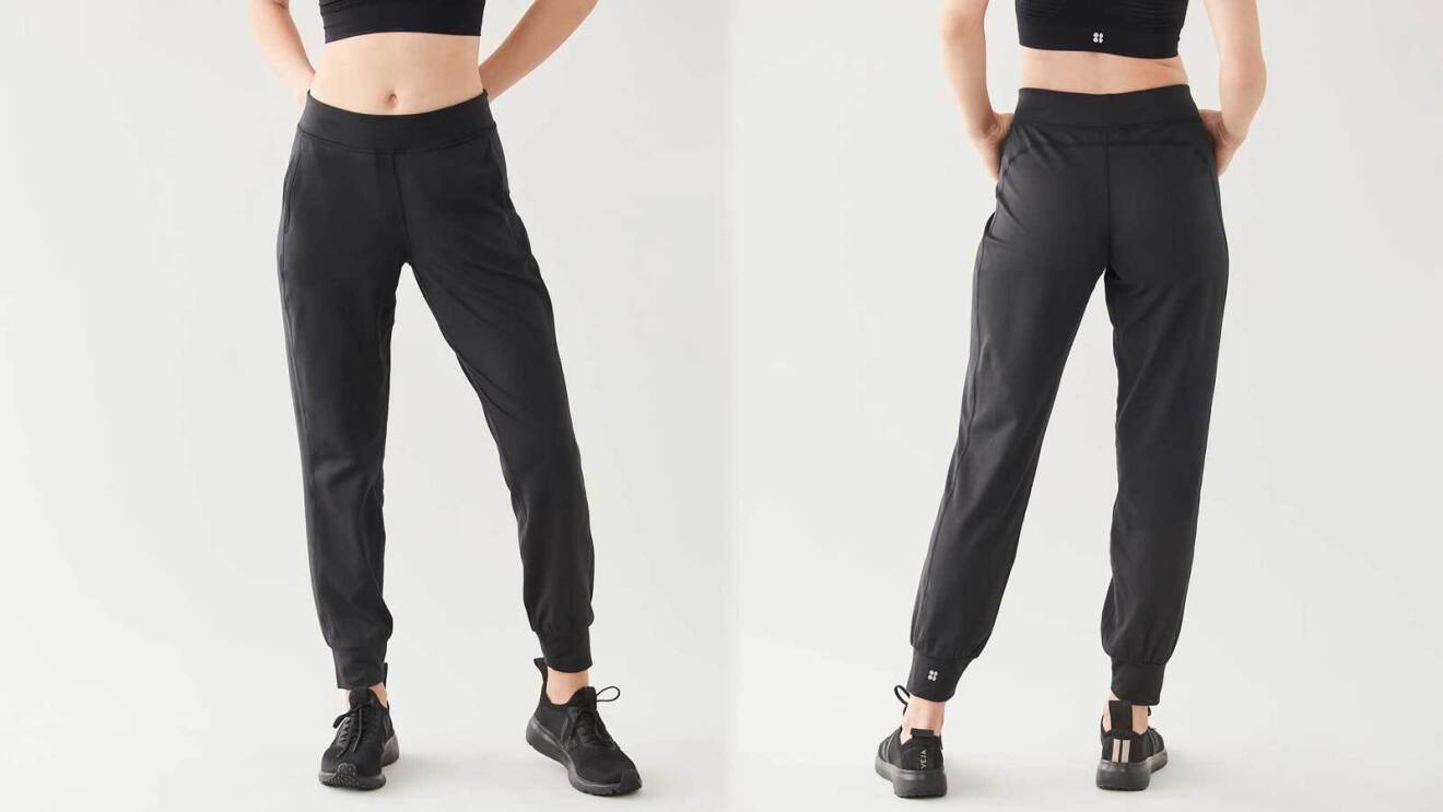 An image of a model wearing black jogger pants from Sweaty Betty. The pants are tight on the ankle and looser around the legs. The waistband hits just below the bellybutton.