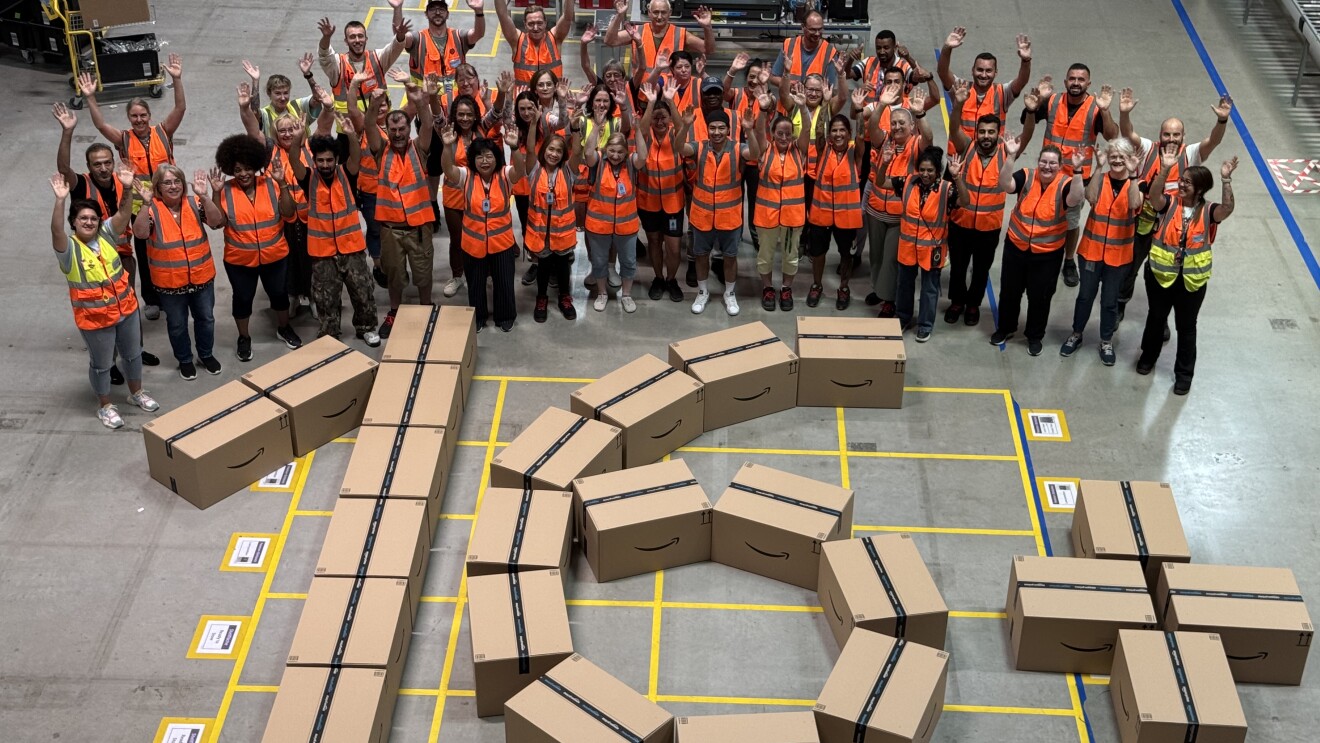 Mitarbeitende stehen im Logistikzentrum um die aus Paketen gelegte Zahl 16+