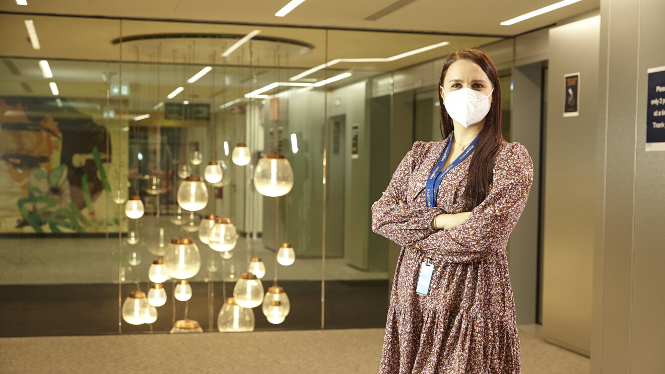 Ragazza ripresa a mezzo busto all'interno degli uffici Amazon. Ha le braccia conserte, indossa un abito colorato e ha una mascherina sul volto.