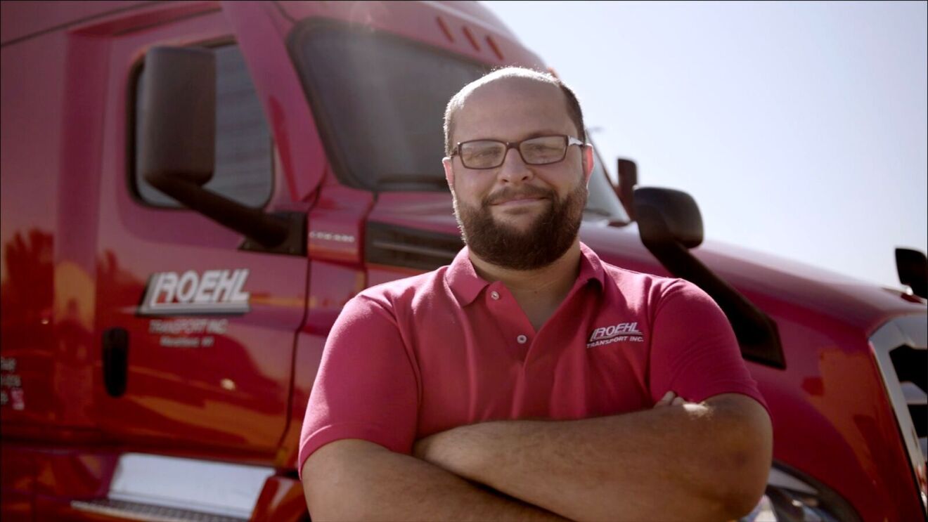 An image of Isaac, dressed in a red shirt, standing in front of a red truck, who works on Amazon's transportation team.