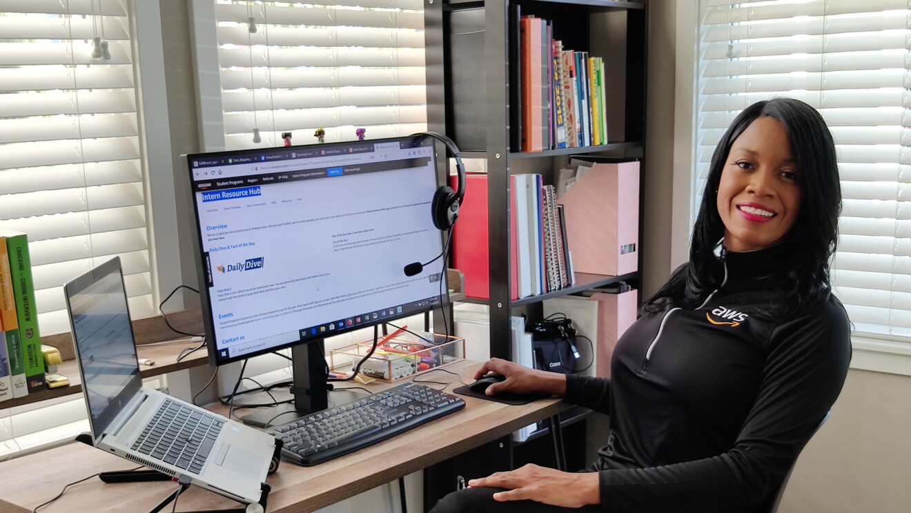 A woman sits at a desk with a laptop and a computer monitor.