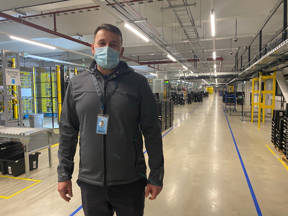 Richard Burtenshaw, FC Associate , he is wearing a face mask and standing in a fulfilment centre.