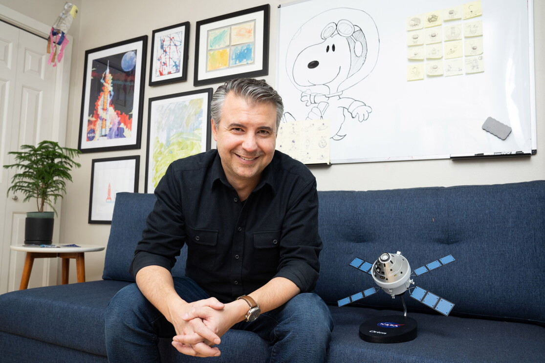 An image of a man smiling for a photo while sitting on a sofa in his office. Next to him is a small satellite device.