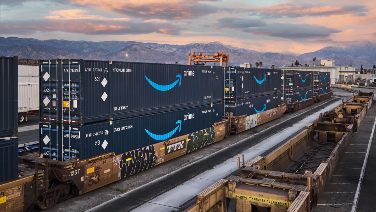 Double-stacked Amazon Prime shipping containers sit on rail cars at an intermodal terminal during sunset with mountains in the background.
