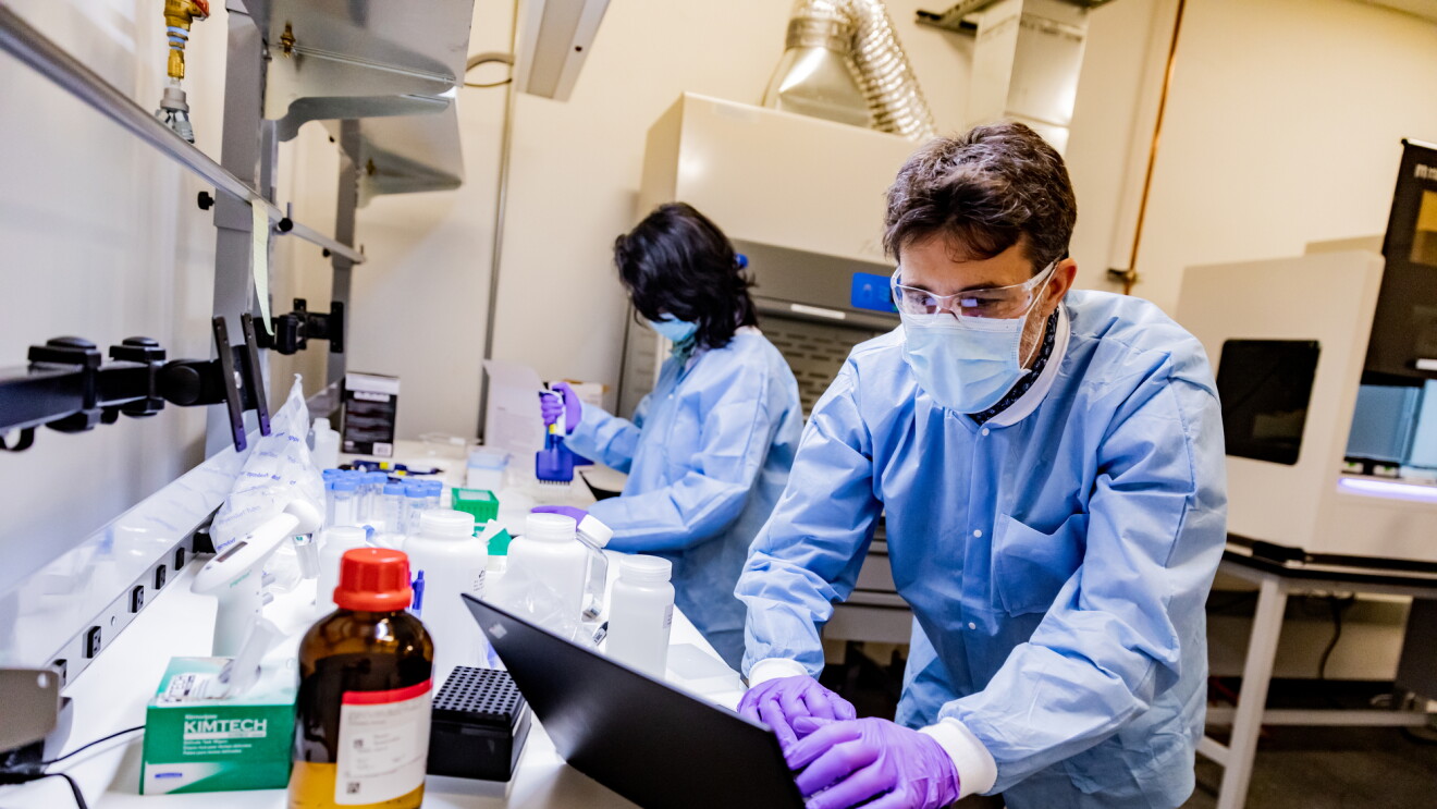 A man in protective gear including safety glasses, mask, gloves, and protective clothing works on a laptop in a laboratory setting. 