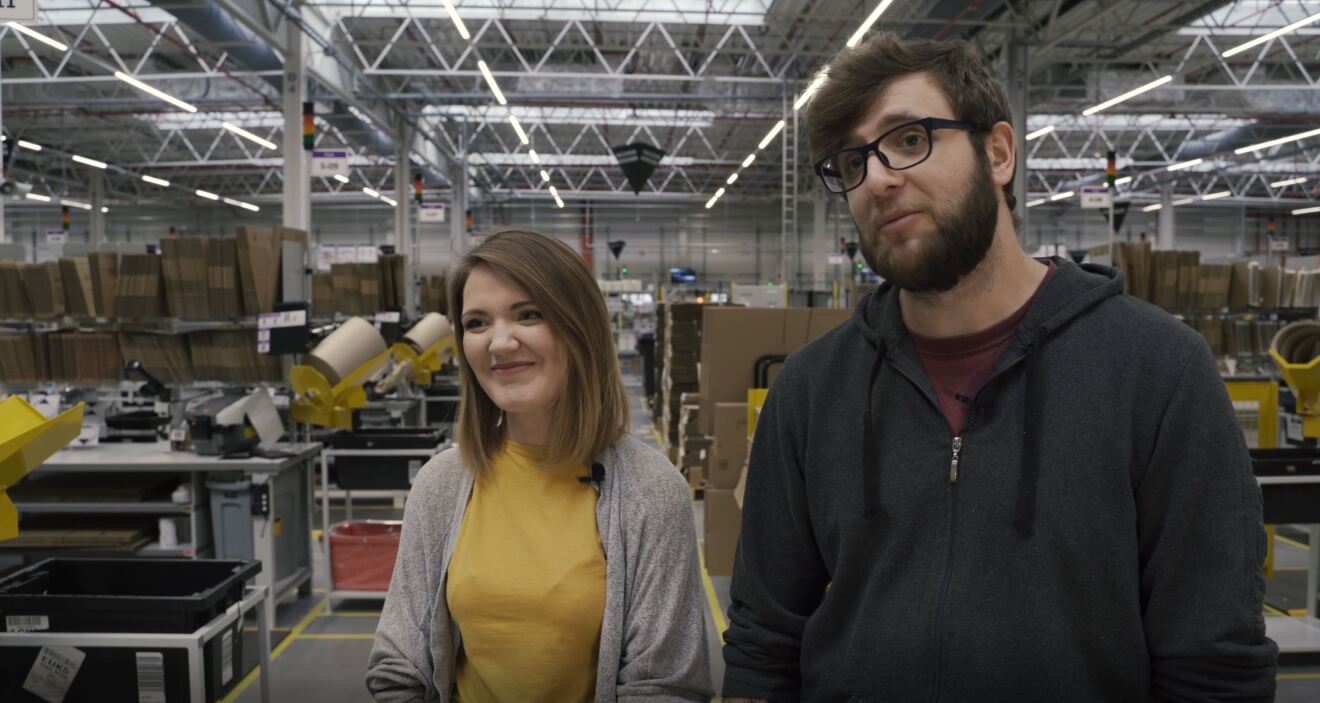 arA woman stands in an Amazon fulfillment center, with a smile on her face. To her left, a man stands. 