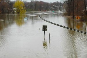 An image of a flooded highway. There is a van floating in the middle. Street signs are partially covered in water with only the signage sticking out. Trees are also sticking out of the water in the background.