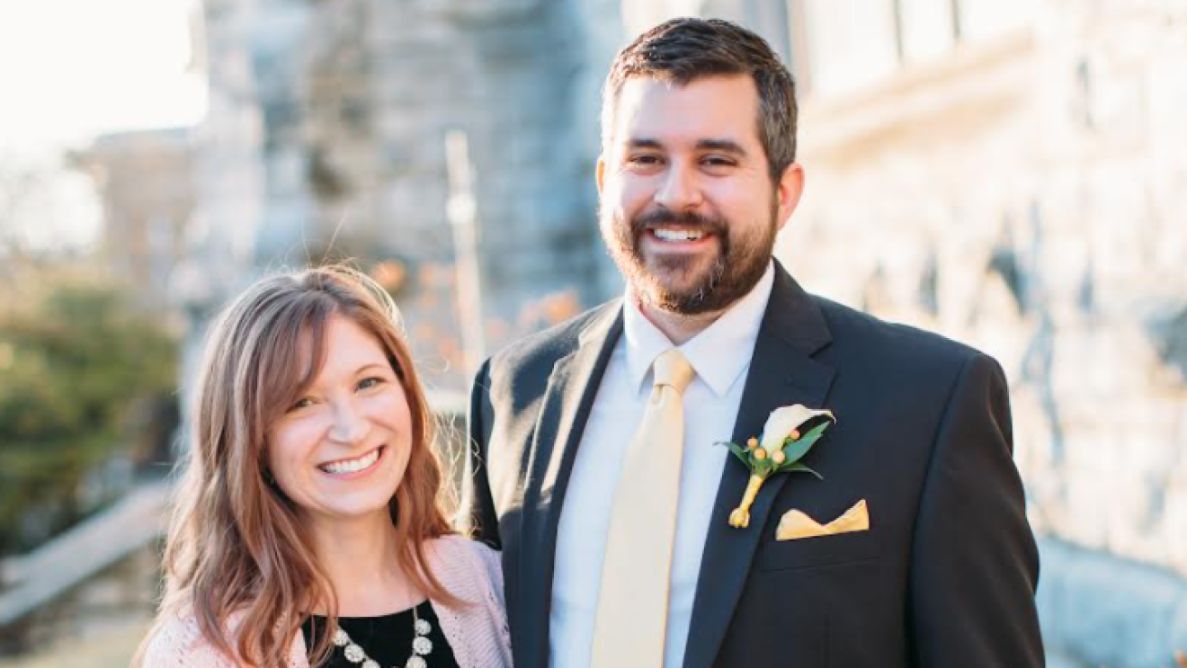 An image of a man and a woman smiling outside of a building while dressed in more formal clothing/