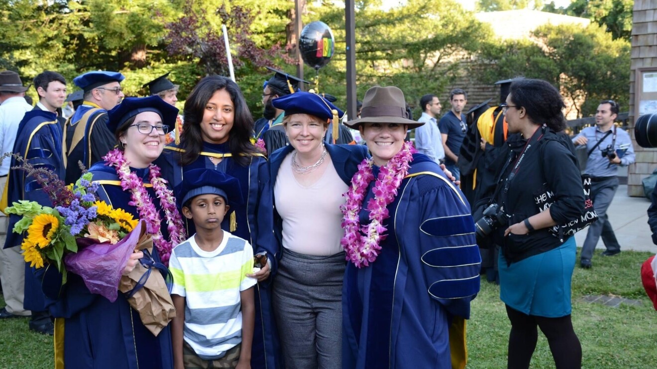 A group of college graduates in caps and gowns smile for a photo with their family members in an outdoor setting.