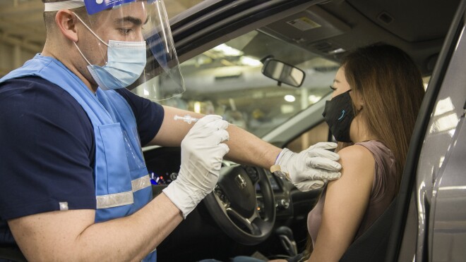 医护人员手持COVID-19疫苗,准备为车内的一名妇女接种疫苗。这名女子戴着一个印有蓝色亚马逊标志的面具。医护人员戴着口罩和面罩。