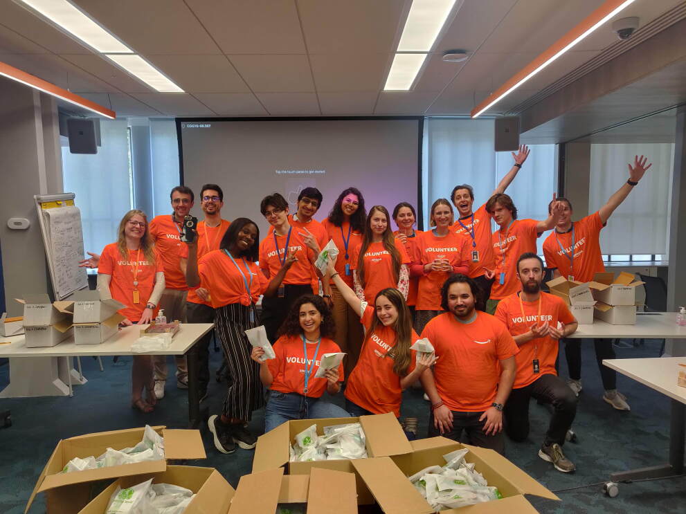 17 personas vestidos con una camiseta naranja con la palabra Volunteer en una sala después de preparar kits para los refugiados. 