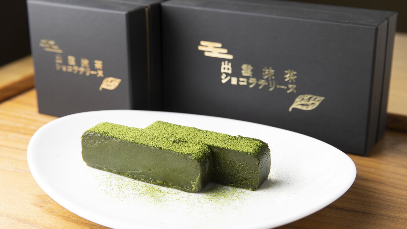 黒い商品箱の前の白い皿の上に置かれた抹茶のスイーツ。生チョコレートのような触感の直方体の上に抹茶が振りかけられている