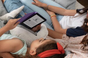 An image of three girls reading on a Kindle device while laying down. The girl holding the device has a stuffed animal next to her and is wearing a red headband. 