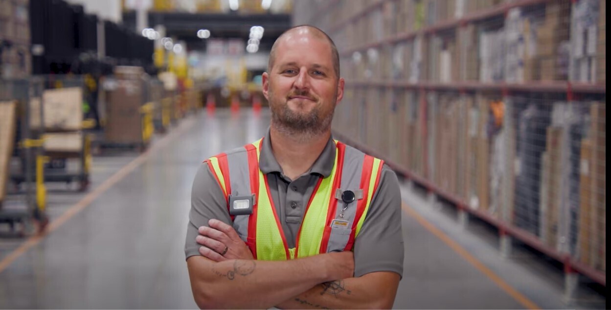 Kevin White wears a safety vest in an Amazon fulfillment center as he crosses his arms and smiles.