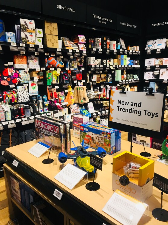 A table of "New and Trending Toys" at the Amazon 4-star store. On the table are a Monopoly board game, LEGO City set, and COZMO robotic toy. Each product has a digital price tag and a customer review displayed alongside the item. Behind the table are Gifts for Pets, Gifts for Grown-Ups, and Candles & Home Decor.