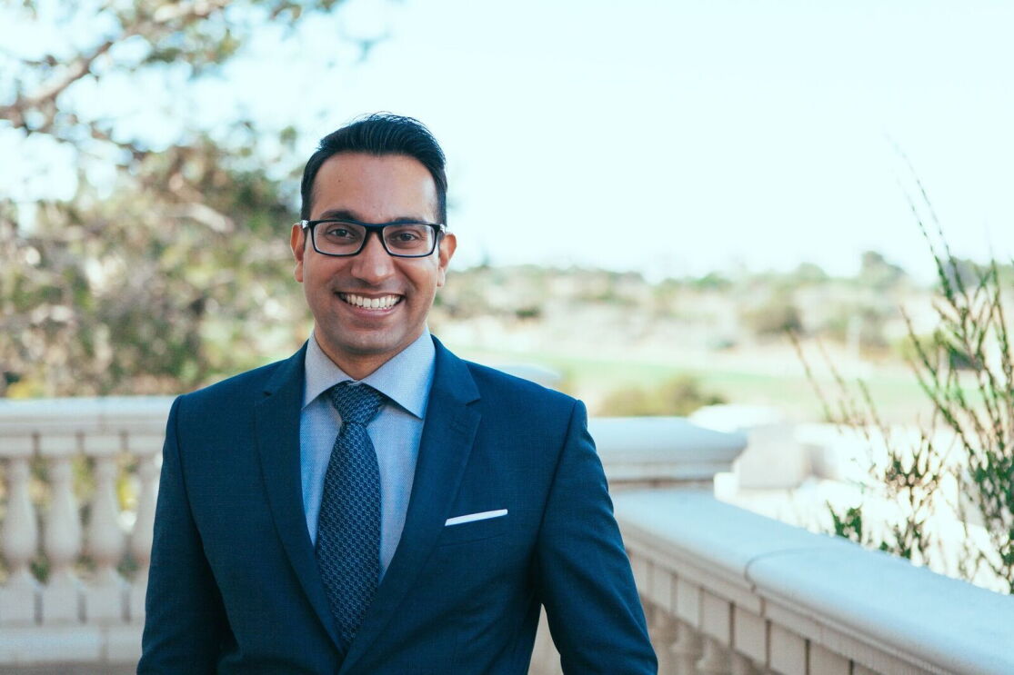 Akash wears a navy suit as he stands outside and smiles for the camera. He has dark hair and wears black-framed eyeglasses.