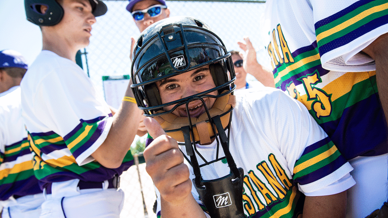 An image of a person wearing a baseball helmet and uniform giving a thumbs up. The background shows other people in the same uniform doing a handshake.