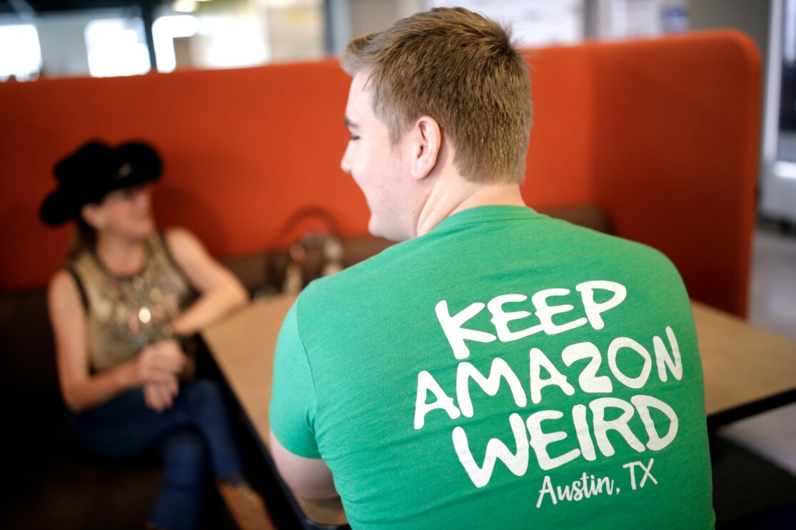 An Amazonian sits, facing away from the camera, in the Austin, TX Amazon office. The back of his shirt says "Keep Amazon weird - Austin, TX." Across the table sits another person, wearing a tank top, jeans, and brimmed hat.