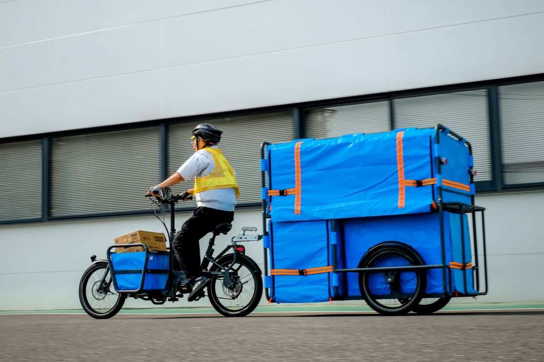 ヘルメットを被って黄色い安全ベストを着ている男性がカゴとリヤカーがついた電動アシスト付き自転車に乗っている。