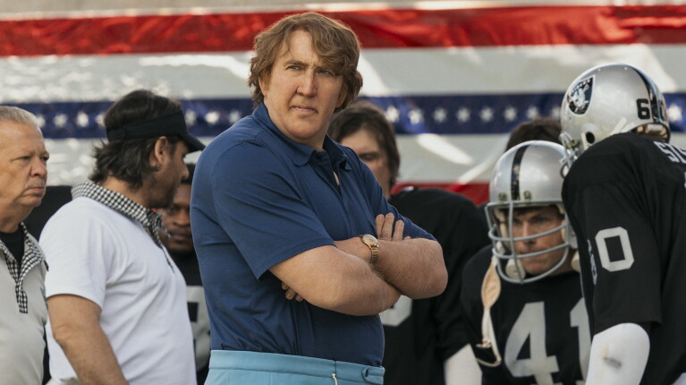 American football coach, Madden, looks out with his players in the background