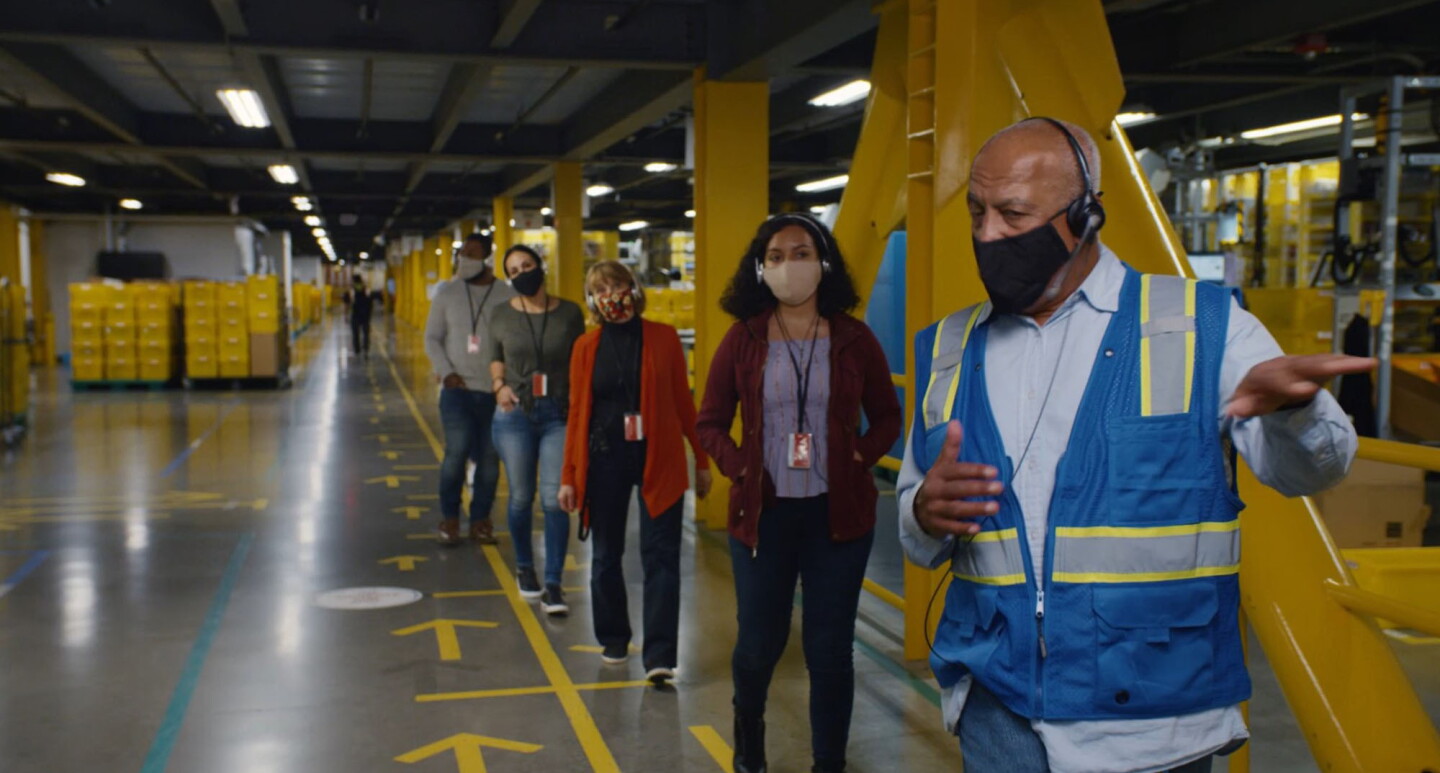 A man wearing a mask leads a line of four masked individuals on a tour at an Amazon fulfillment center. 