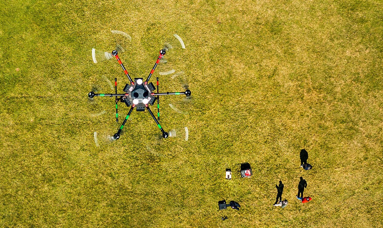 An image of a drone flying over a green field. People below are controlling it.