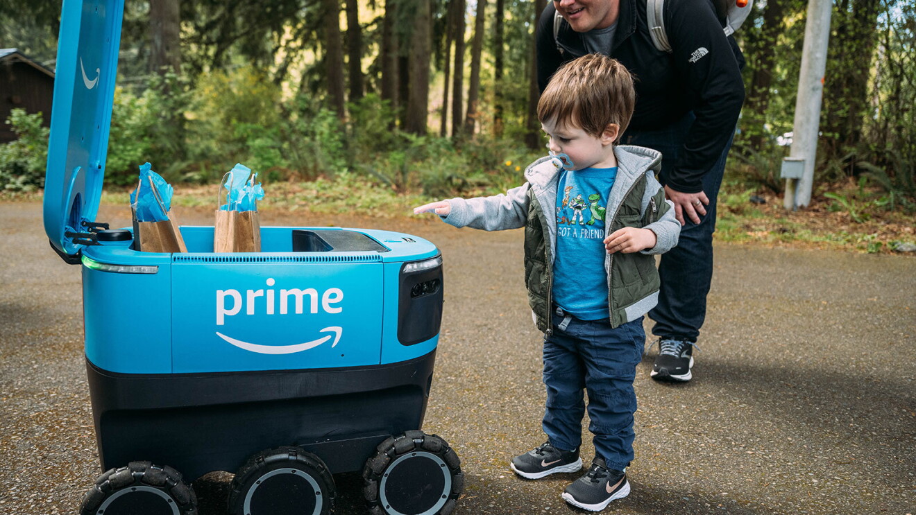 A young boy reaches toward a Scout delivery robot. His father smiles as he leans toward him. 