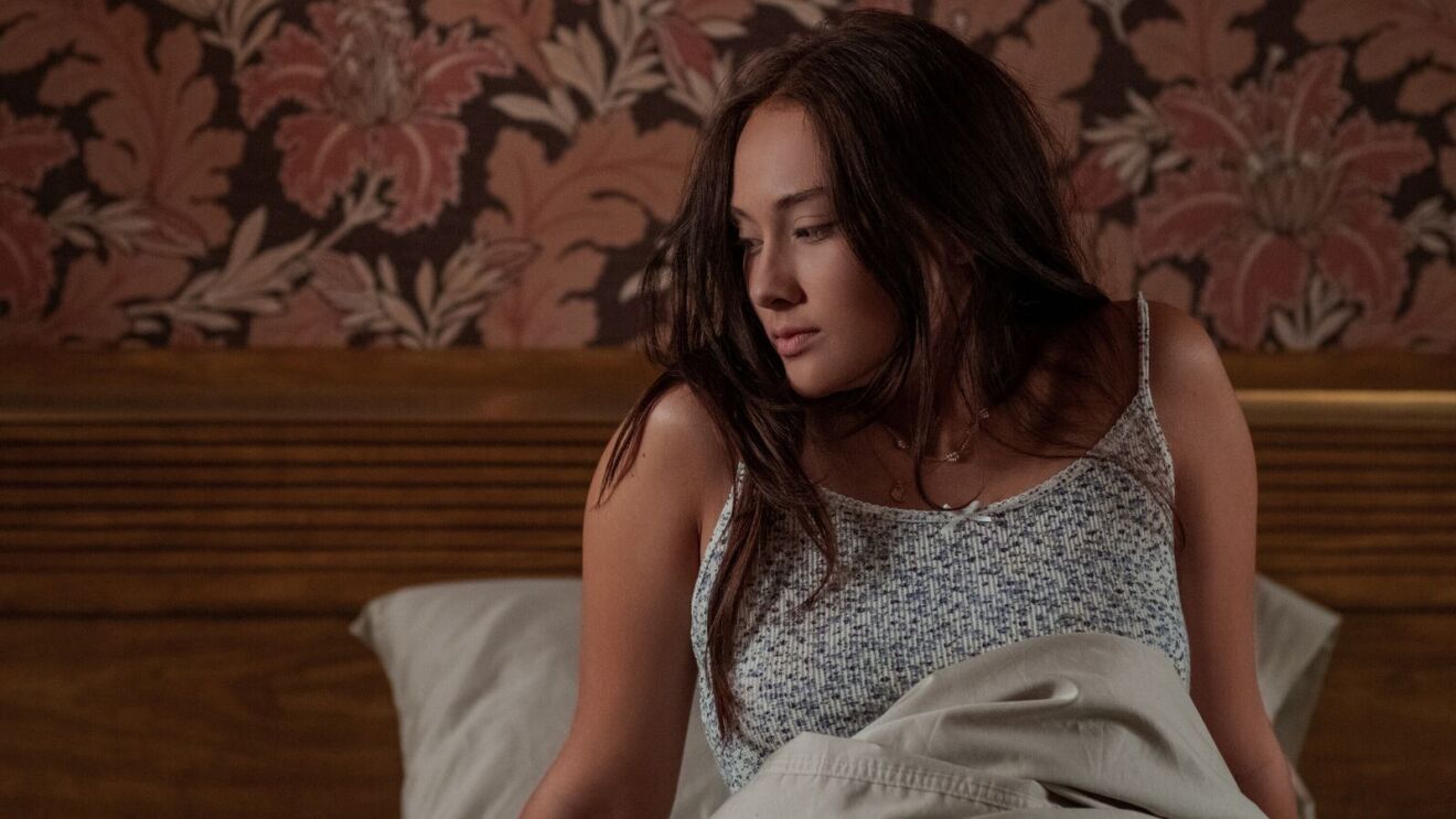 Belly (Lola Tung) in white floral tank top on bed in dimly lit room