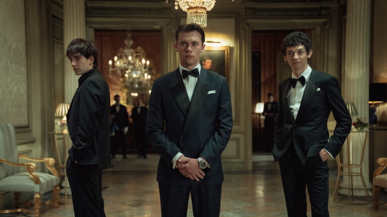 Three young men in formal tuxedos standing in an opulent mansion hallway