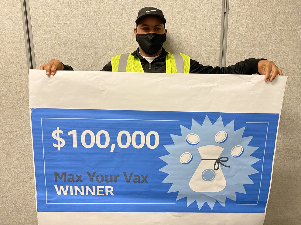 An image of a man holding a large check that says "$10,000 Max Your Vax WINNER"