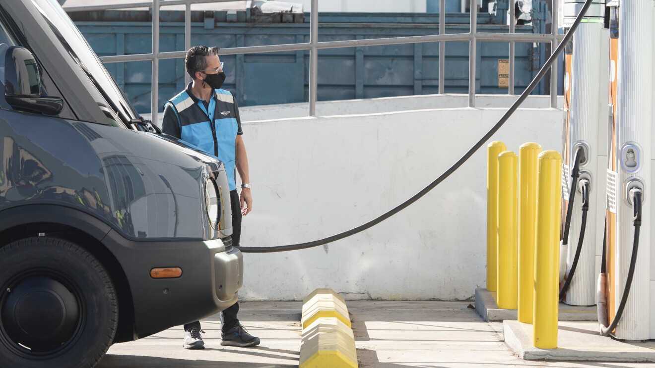 An Amazon-branded Rivian van gets charged at an electric station by a man wearing a face mask during the pandemic.