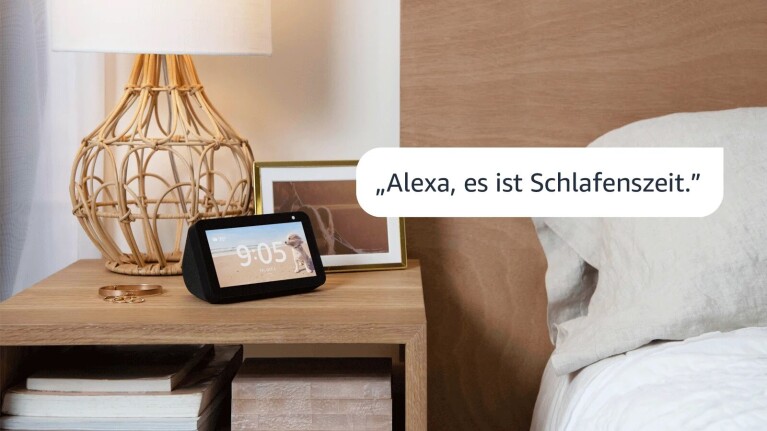 Ein Bild eines Amazon Echo Show-Geräts, das auf einem Nachttisch aus Holz neben einem Bett steht. Neben dem Gerät befinden sich ein Bilderrahmen, eine Lampe und Schmuck.