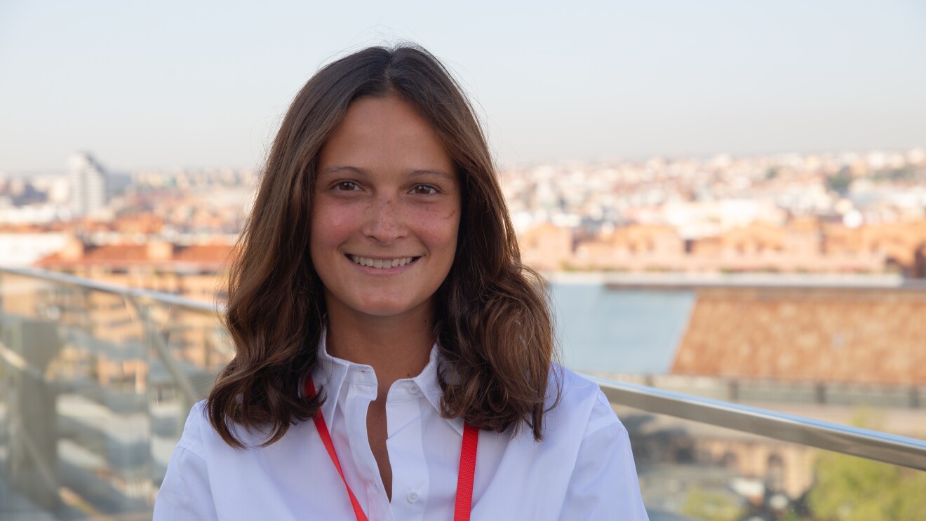 María Robert, Senior Program Manager de Experiencia del Cliente. De fondo el paisaje de Madrid. Está en el último piso de las oficinas corporativas. Hay una barandilla de color metal. María tiene el pelo castaño un poco más allá del hombro. Lo lleva suelto. Está morena, tiene pecas y lleva una camisa blanca.