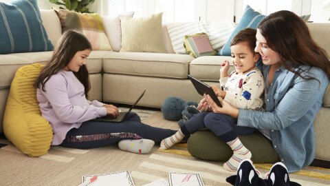 一位母亲带着两个孩子坐在客厅的地板上使用他们的电脑和平板电脑。