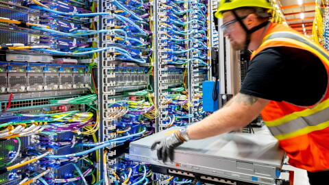 A man in an orange safety shirt and yellow safety hat in a data centre, working with data centre tower parts. 