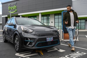 An image of a man walking around an electric car with a cord that he is going to plug in to charge it. The car and the man are in the parking lot in front of an Amazon Fresh store.