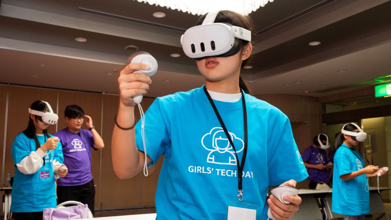 青いTシャツを着ている女性がVRヘッドセットをかけてコントローラーで動いているようだ。彼女の後ろには他の女性3人が同じことをしている。
