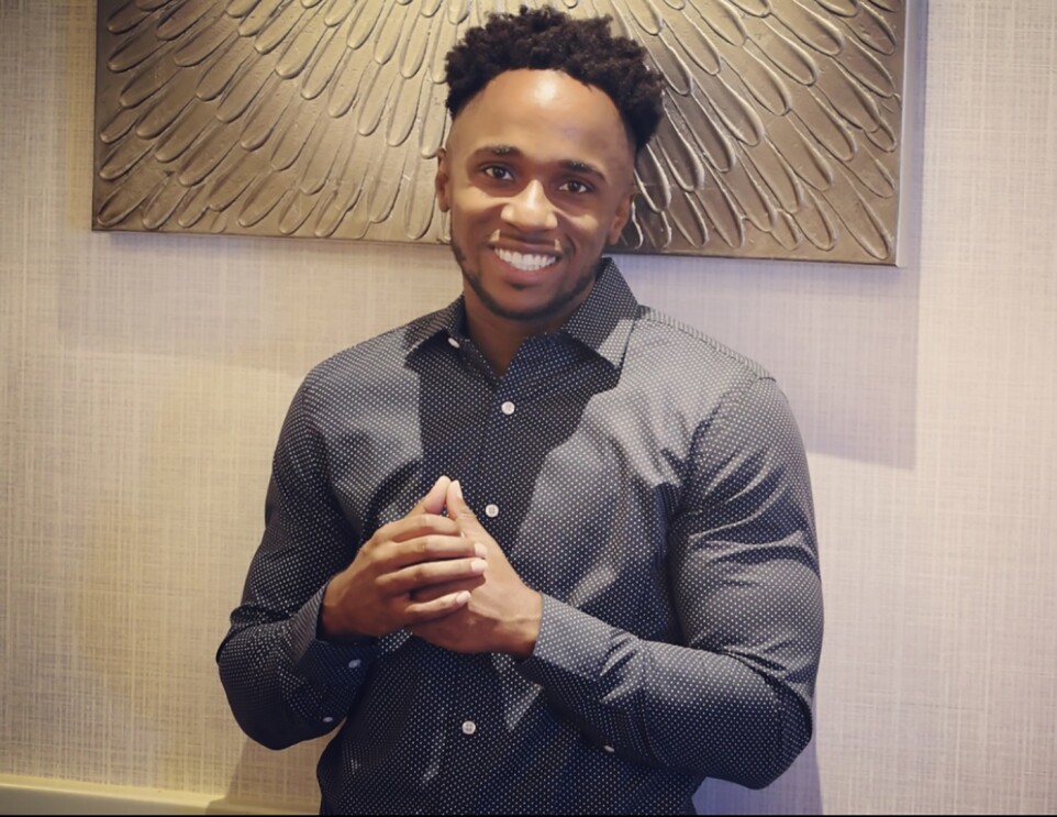 A Black man wearing a button up shirt smiles at the camera. 