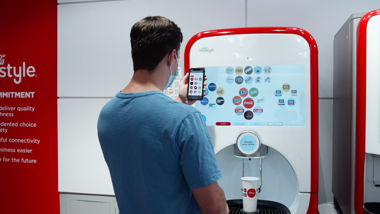 A man wearing a mask and using his mobile phone stands before a Coca-Cola Freestyle touchless experience built on AWS to make his drink selection.