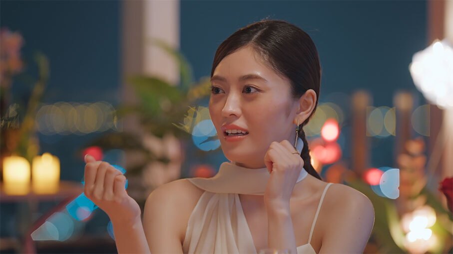 夜景が見える窓を背景に、女性のクローズアップ。白いドレスを着用、身振りを交えて話をしている。背後には植物やキャンドル。