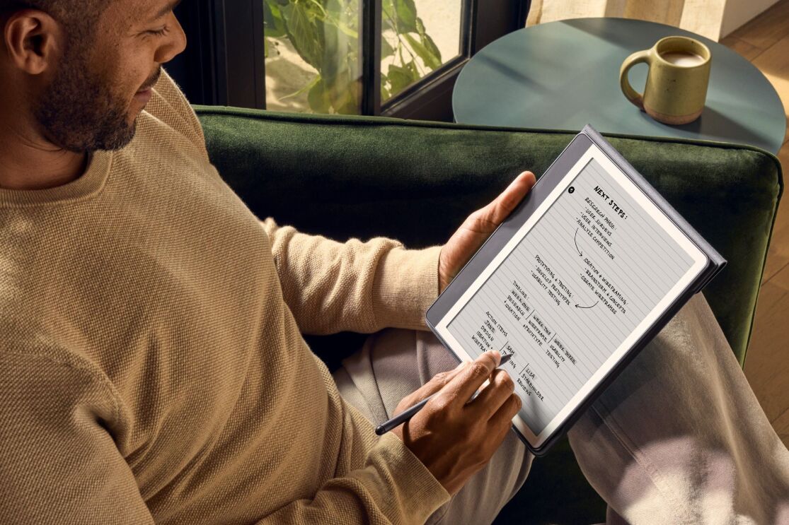 茶色のセーターを着ている男性がタブレットで文章を書いている。彼は明るい日差しが差し込むリビングの緑色のソファに座っている。その隣のコーヒーテーブルにマグカップが置かれている