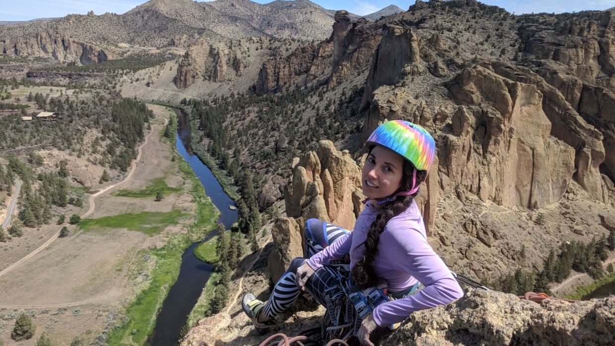 Marta Fraga en la cima de una montaña mirando a cámara. Va vestida con ropa de escalada de color morado y un casco también de varios colores dorados. Desde la cima, sentada y con unas cuerdas en un lado mira a cámara. De fondo un río que cruza la imagen y montañas rocosas sin mucha vegetación.