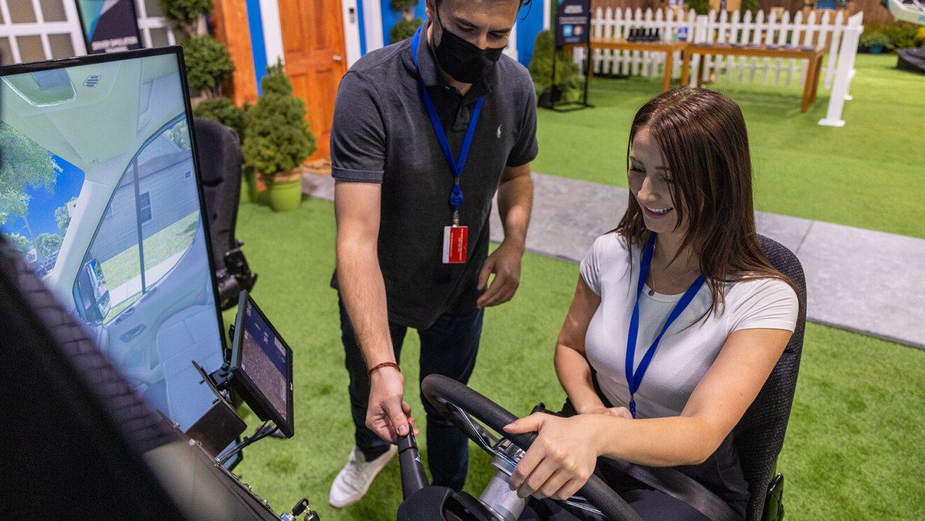 An image of a woman sitting in the driver's seat of an automated driving system. A man behind her is pointing to the windshield wipers to show her where they are.