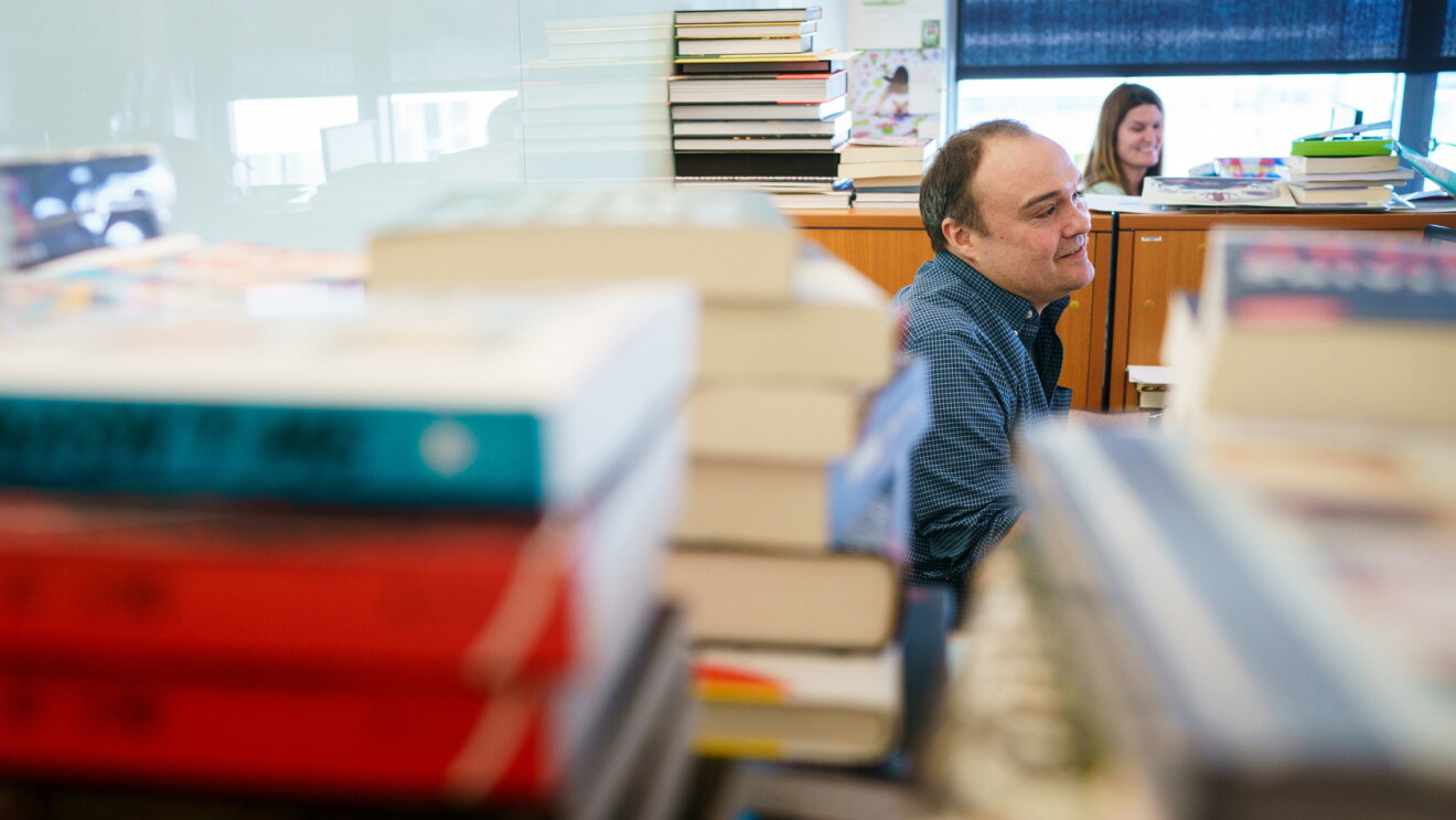 A seated man is mostly obstructed by stacks of books in the foreground of this image. He wears a dark button-down shirt and has brown hair. In the background is a woman with long brown hair as well as more stacked books on top of book shelves.