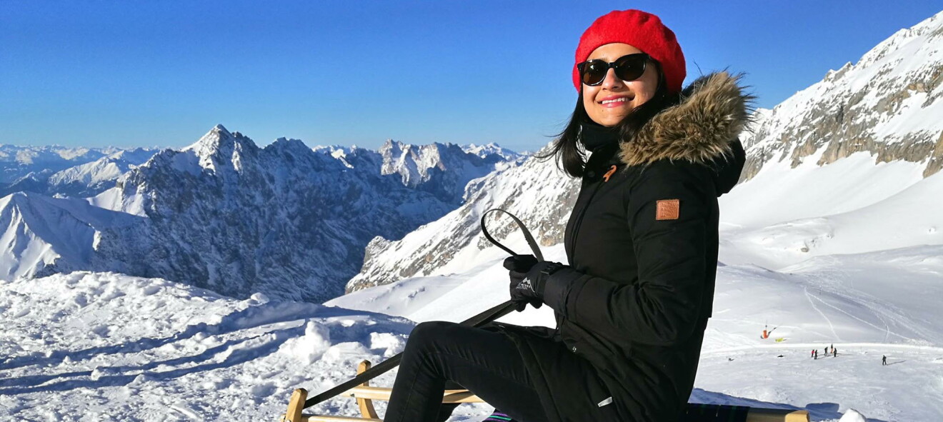 Mehak in den Alpen beim Schlitten fahren. Sie trägt eine rote Pudelmütze und eine Sonnenbrille. Im Hintergrund sieht man die schneebedeckten Alpen.